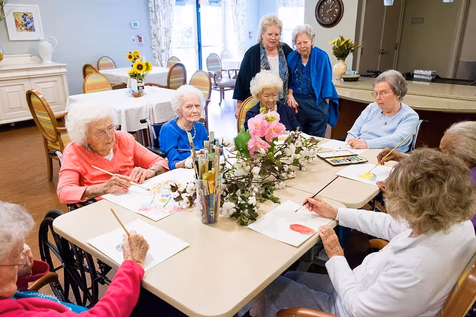 A group of elderly women seated around a table engaged in painting and drawing activities. Two women stand behind them observing. The table has art supplies and a floral centerpiece. The room has several tables and chairs with a bright, welcoming atmosphere.