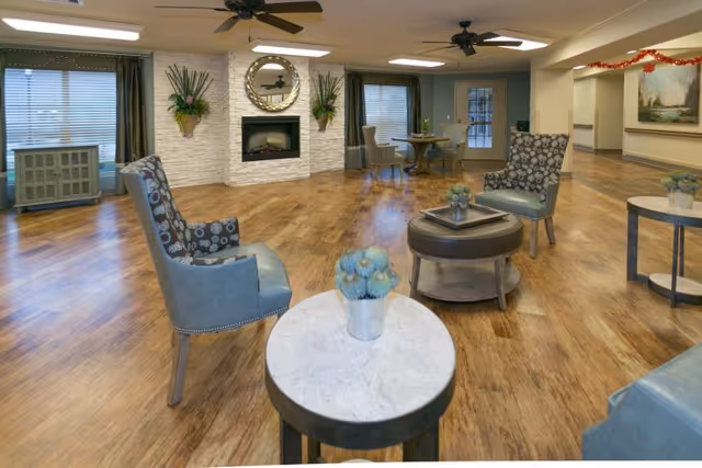 A spacious living room area with wooden flooring, several upholstered chairs with patterned fabric, small round tables with decorative plants, a white stone fireplace with a round mirror above it, and large windows with blinds. Ceiling fans and recessed lighting are visible on the ceiling. There is a table with chairs near the back and a doorway leading to another room.