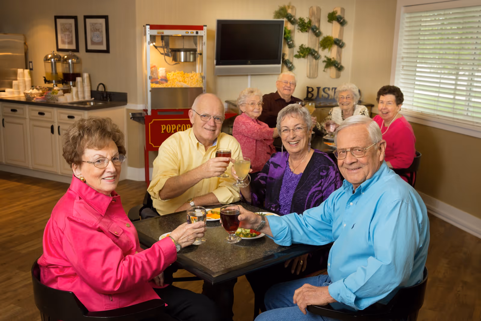 A group of elderly people sitting around a dining table in a senior living facility, smiling and raising their glasses in a toast. The room has a popcorn machine, a TV mounted on the wall, and a counter with cups and juice dispensers in the background.