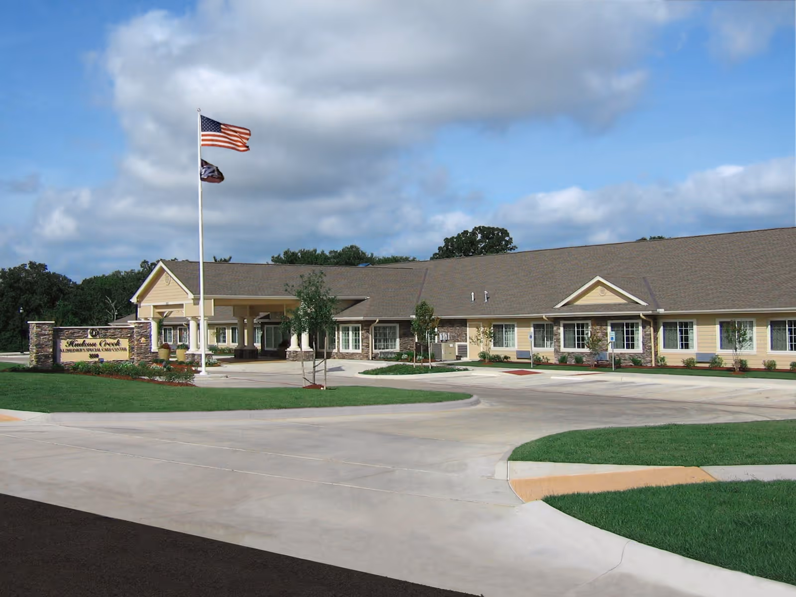 Exterior view of Hudson Creek Alzheimer's Special Care Center building with a driveway, landscaped lawn, and two flagpoles displaying the American flag and another flag under a partly cloudy sky.