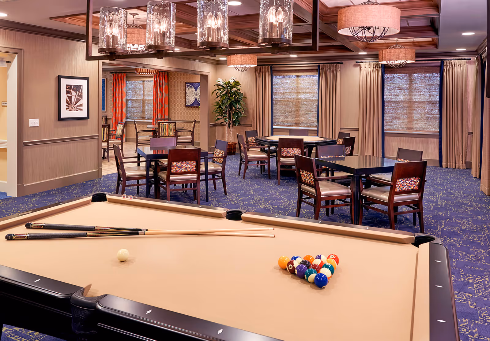 Interior view of a recreational room featuring a beige pool table with two cues and a rack of billiard balls in the foreground. Behind the pool table, there are several tables and chairs arranged for social or gaming activities. The room has blue patterned carpet, beige walls, large windows with curtains, and multiple ceiling light fixtures.