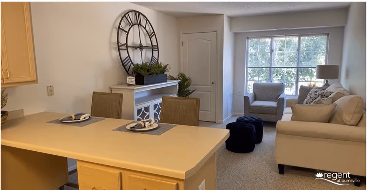 A cozy living area with a beige sofa, an armchair, two dark blue ottomans, and a large window letting in natural light. In the foreground, there is a kitchen counter with two place settings, and a white cabinet with a large decorative wall clock and a plant on top.