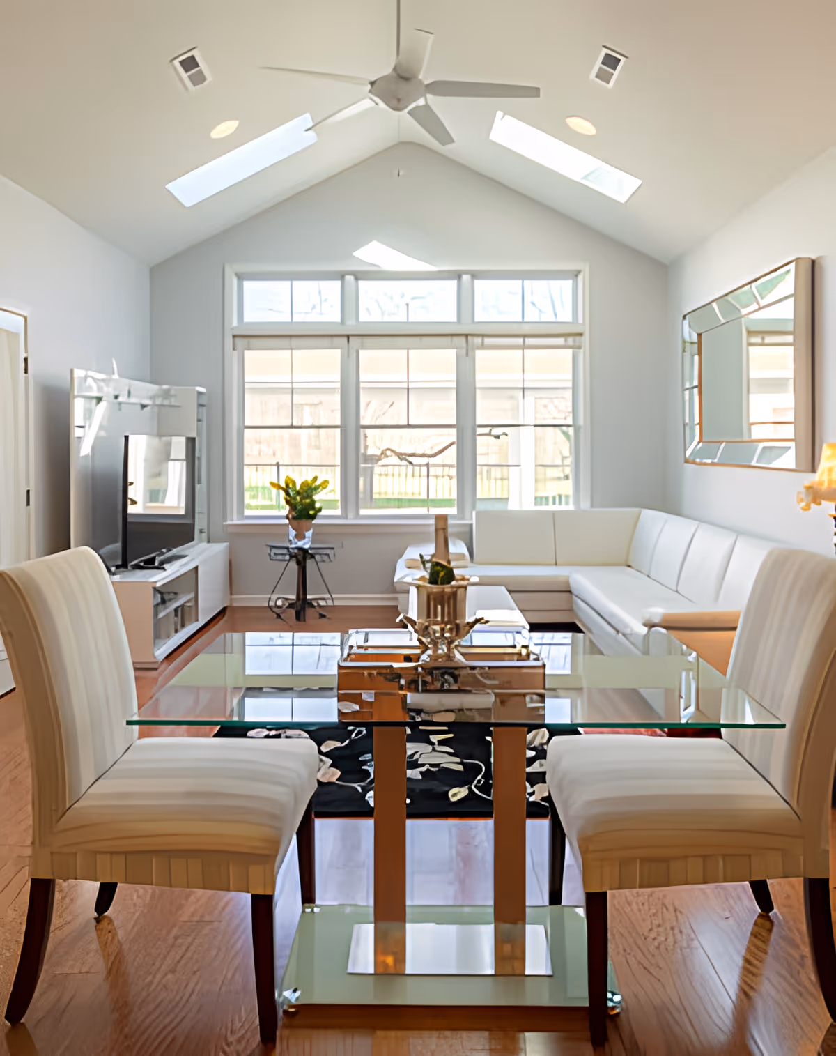 Bright living room with a glass dining table and four upholstered chairs in the foreground. A white sectional sofa is placed against the far wall under a large window with multiple panes. The room has a high vaulted ceiling with skylights and a ceiling fan. A large mirror hangs on the right wall, and a TV is positioned on a stand to the left. The floor is wooden, and a decorative rug is partially visible under the table.