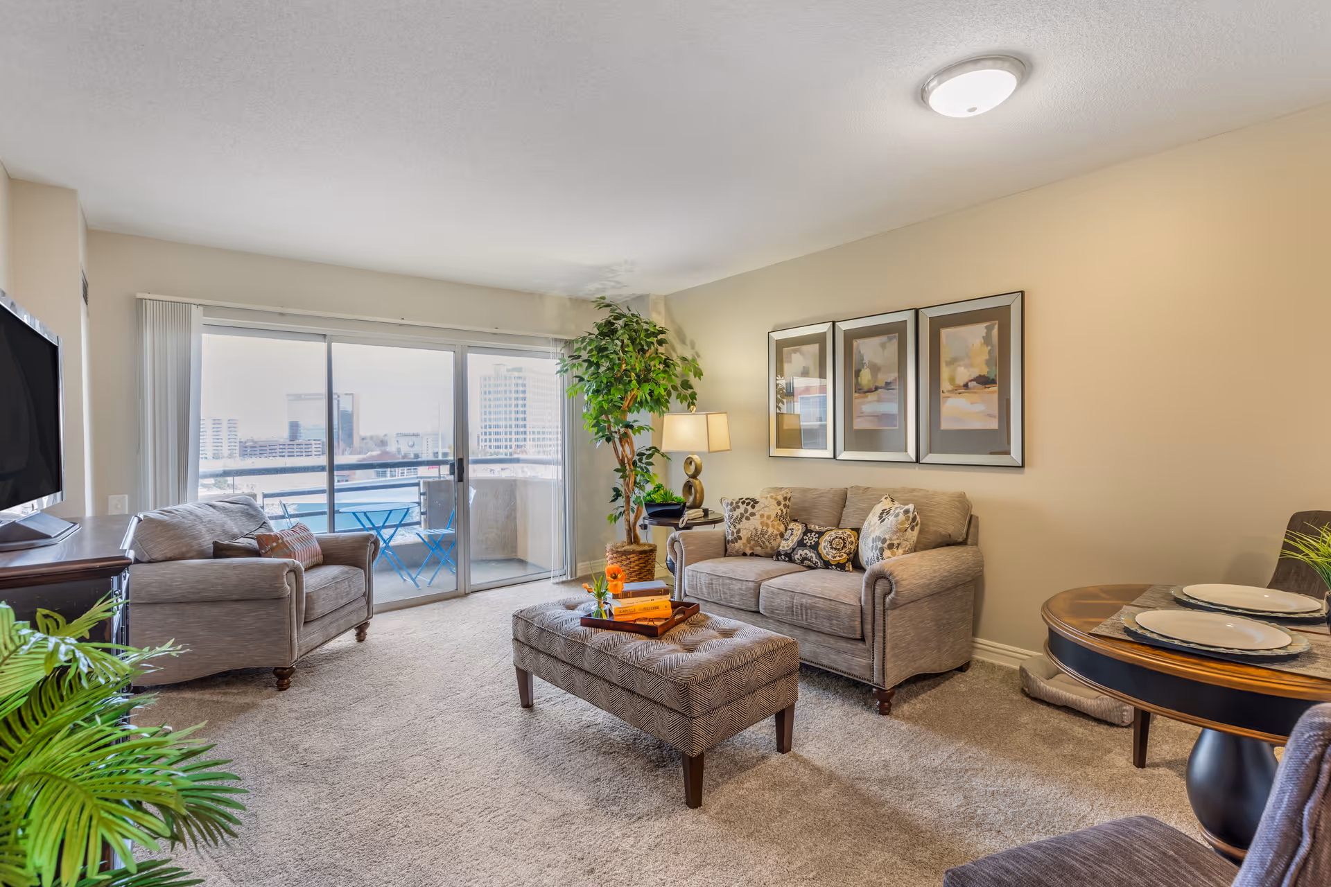 A cozy living room with beige walls and carpeted floor. The room features a gray sofa and matching armchair with decorative pillows, a patterned ottoman with a tray holding books and a small flower vase, a round dining table set with plates and placemats, a tall potted plant, and a sliding glass door leading to a balcony with a small blue table and chairs. Three framed abstract paintings hang on the wall above the sofa.