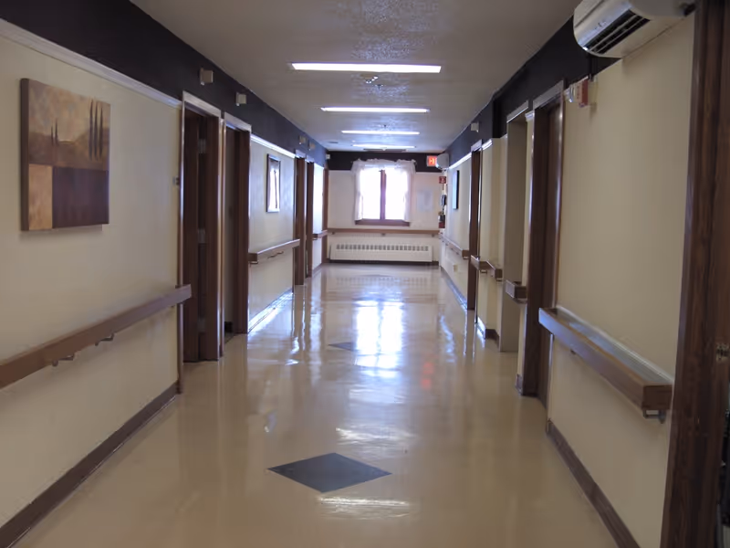 A clean, empty hallway in a healthcare facility with beige walls, wooden handrails on both sides, several doorways, a window at the end with curtains, and overhead fluorescent lighting.