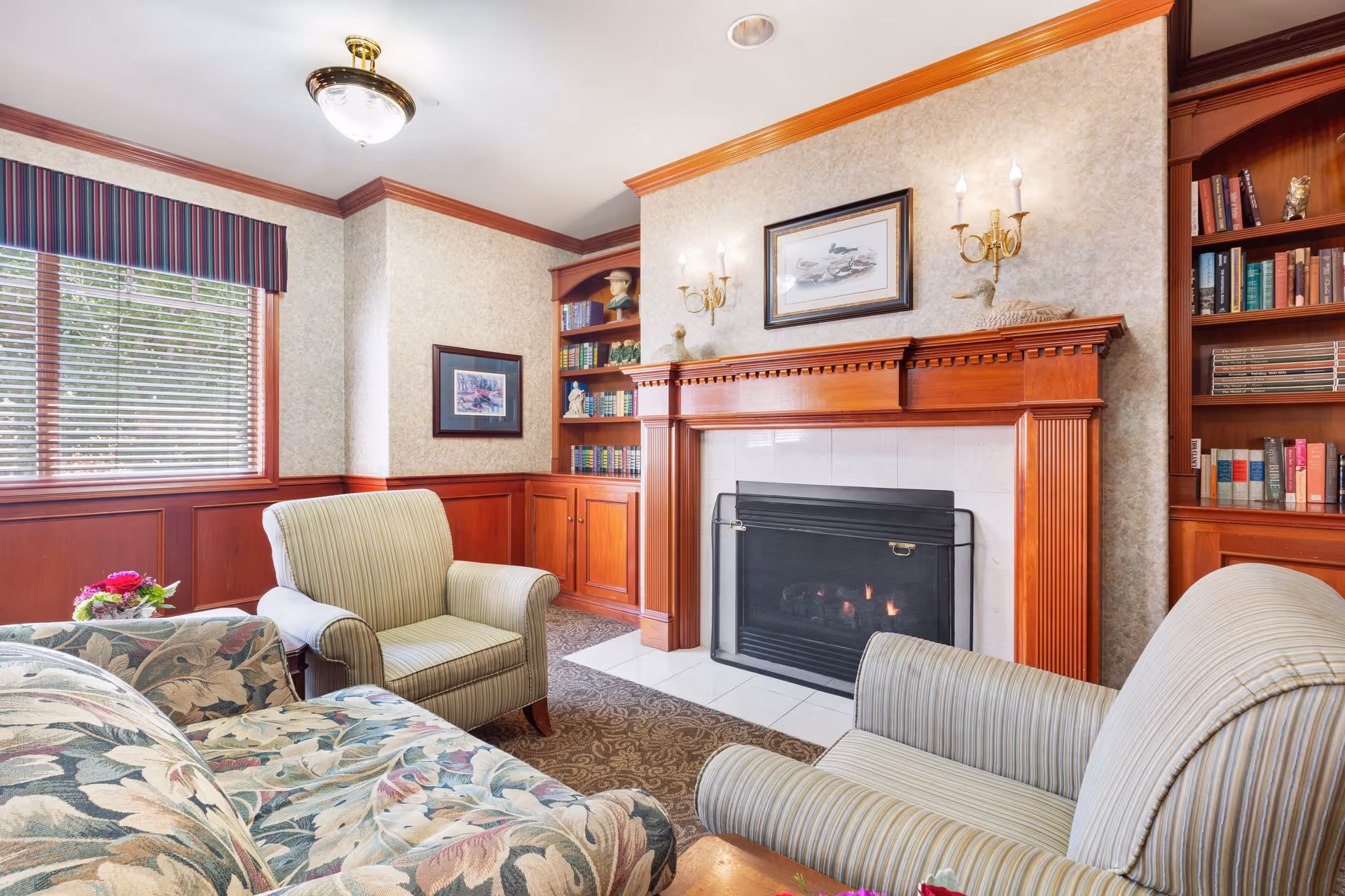 A cozy living room area with a floral patterned sofa and two striped armchairs arranged around a fireplace with a wooden mantel. The room features built-in wooden bookshelves filled with books and decorative items, a window with blinds and a striped valance, and framed artwork on the walls. The lighting includes a ceiling fixture and two wall sconces above the fireplace.
