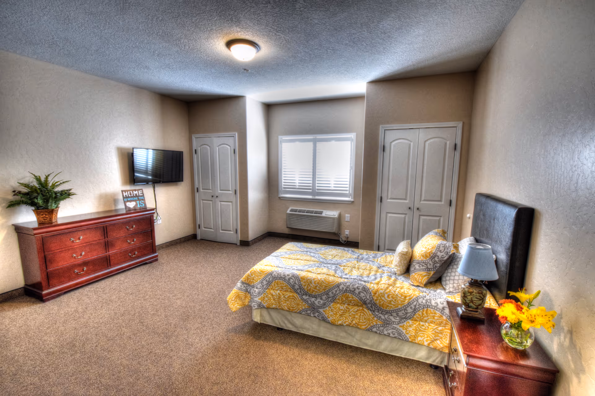 A bedroom with a bed covered in a yellow and gray patterned quilt, a wooden nightstand with a lamp and a vase of yellow flowers, a wooden dresser with a potted plant and a small sign that reads 'HOME is where the heart is', a wall-mounted flat screen TV, two closed closets, a window with blinds, and beige walls and carpet.