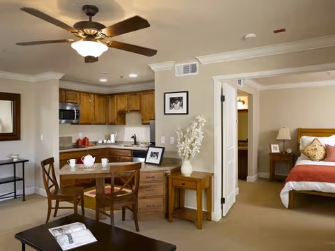 Open-plan apartment living area with a ceiling fan, small dining table and kitchenette, and a bedroom visible through an open doorway.