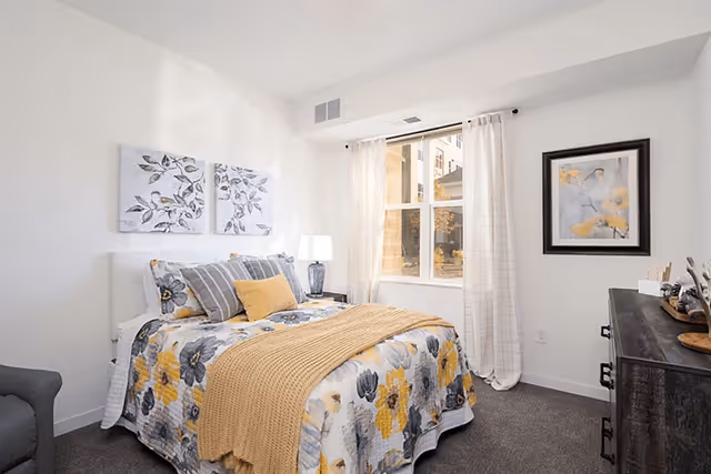A bright and cozy bedroom with a bed dressed in floral bedding featuring yellow and gray flowers, complemented by a mustard yellow throw blanket and multiple pillows. The room has white walls with two floral paintings above the bed and a framed artwork on the adjacent wall. A window with sheer white curtains lets in natural light. There is a dark wooden dresser on the right side and a gray armchair on the left.