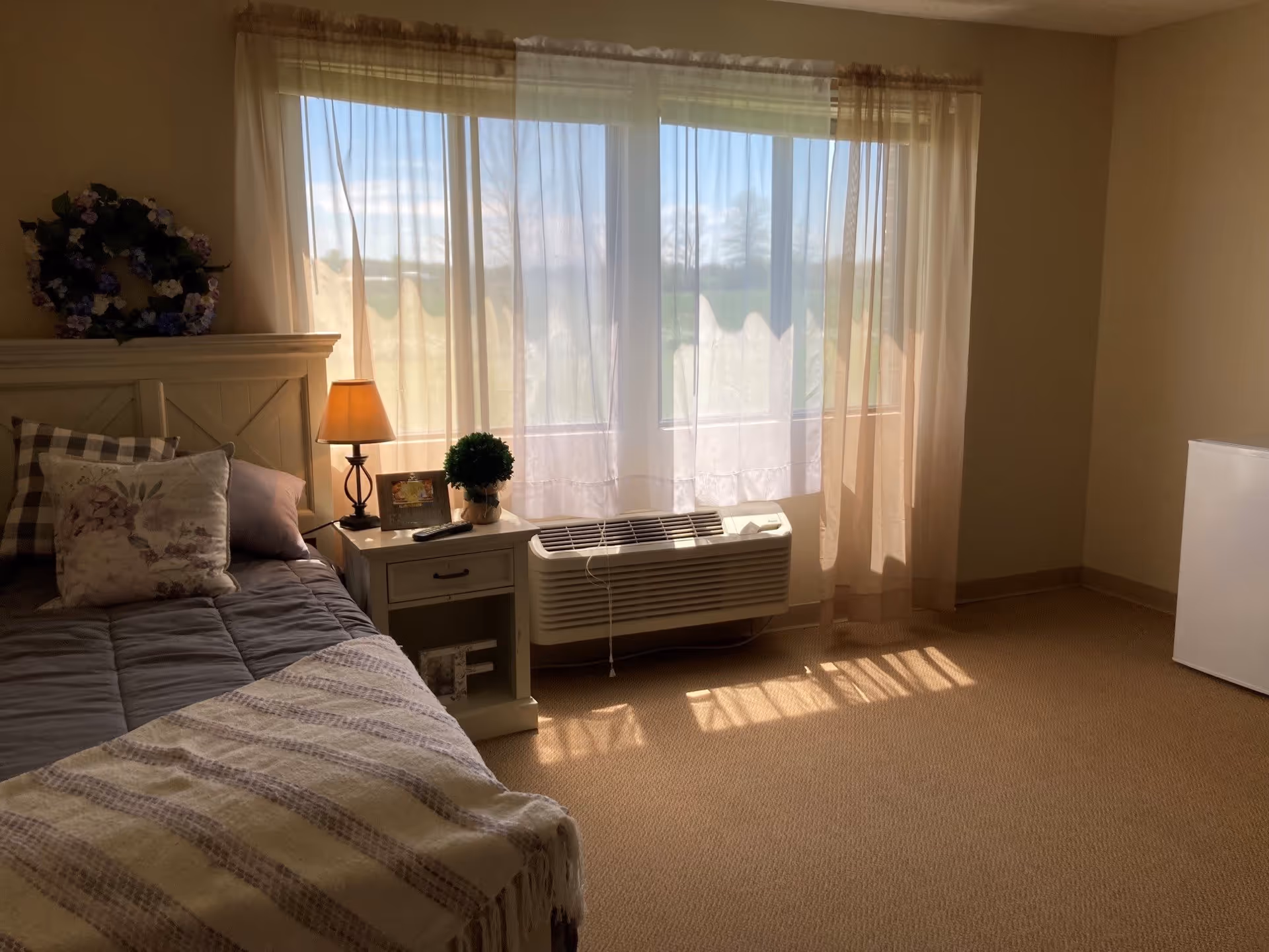A cozy bedroom with a bed on the left side featuring a gray comforter, floral and plaid pillows, and a beige throw blanket. Next to the bed is a white nightstand with a lamp, a small potted plant, and a framed photo. Behind the nightstand is a large window with sheer curtains allowing sunlight to fill the room. Below the window is a wall-mounted air conditioning unit. On the right side of the room, there is a small white refrigerator.
