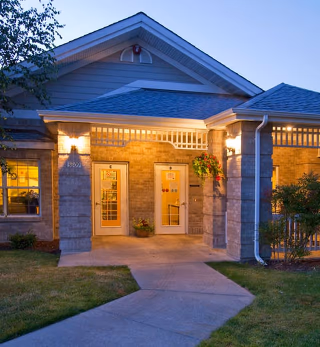 Exterior view of the entrance to Ashley Gardens of Mt. Vernon at dusk, showing a covered porch with two glass doors labeled C and D, illuminated wall lights, hanging flower basket, and surrounding greenery.