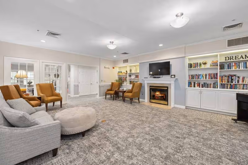 A spacious and well-lit living room area in a senior living facility featuring a gray sofa with cushions, a round beige ottoman, and several mustard-colored armchairs arranged around small tables. The room has a patterned carpet, a fireplace with a TV mounted above it, and built-in bookshelves filled with books and decorative items. The walls are painted light beige, and there are ceiling lights providing illumination.