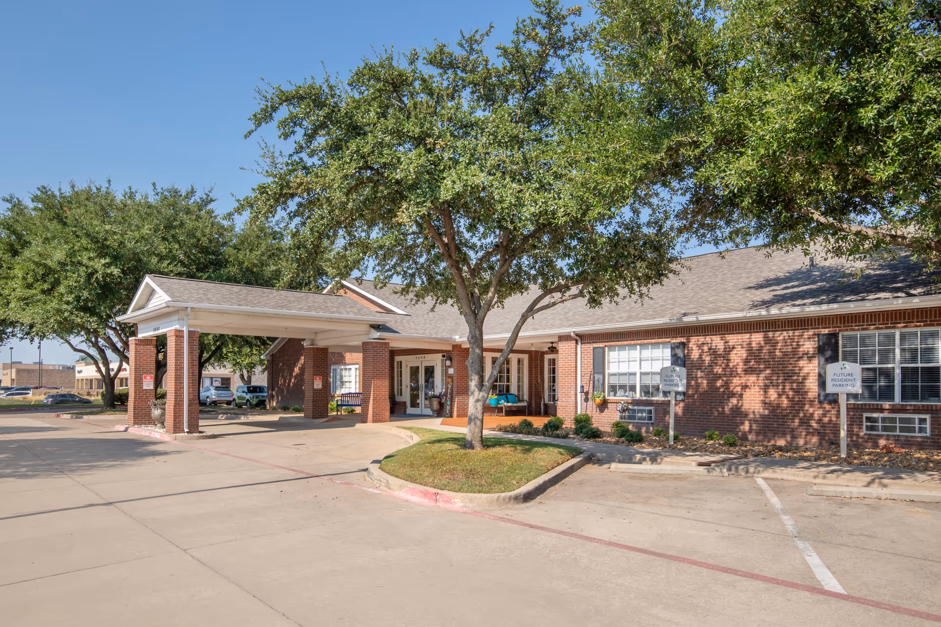Exterior view of a single-story brick building with a covered entrance driveway, surrounded by trees and parking spaces labeled for future resident parking, under a clear blue sky.