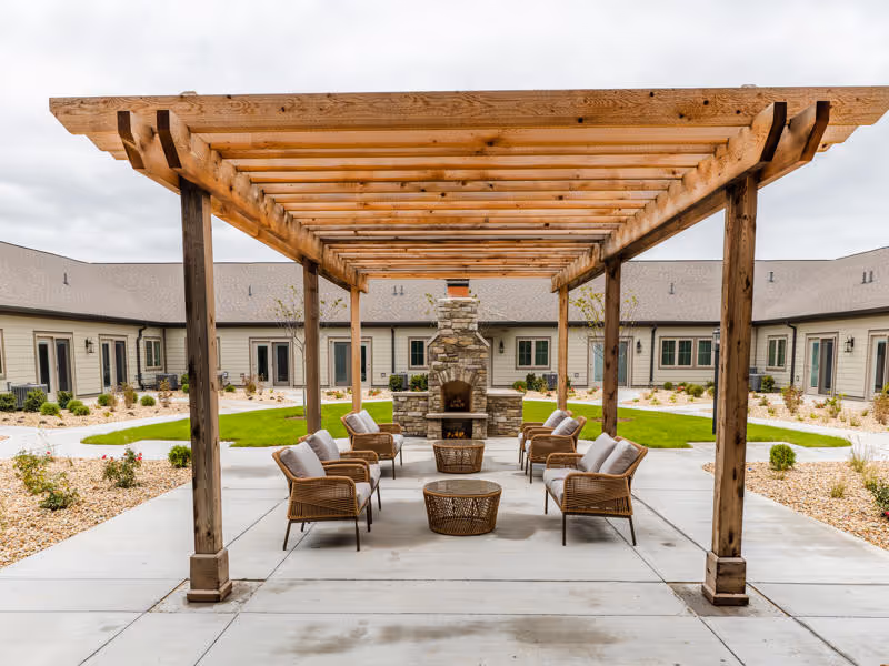 Outdoor seating area at Cedarhurst Senior Living of Frankfort featuring a wooden pergola, cushioned wicker chairs arranged around a stone fireplace, with a courtyard and building wings in the background under a cloudy sky.