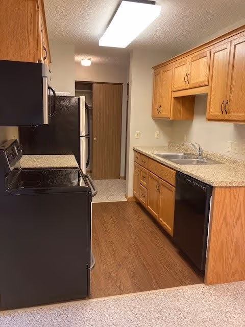 A narrow kitchen with wooden cabinets, a black electric stove and oven, a black dishwasher, a double sink, and a refrigerator. The floor is wood laminate, and there is a bright rectangular ceiling light. A doorway is visible at the end of the kitchen.