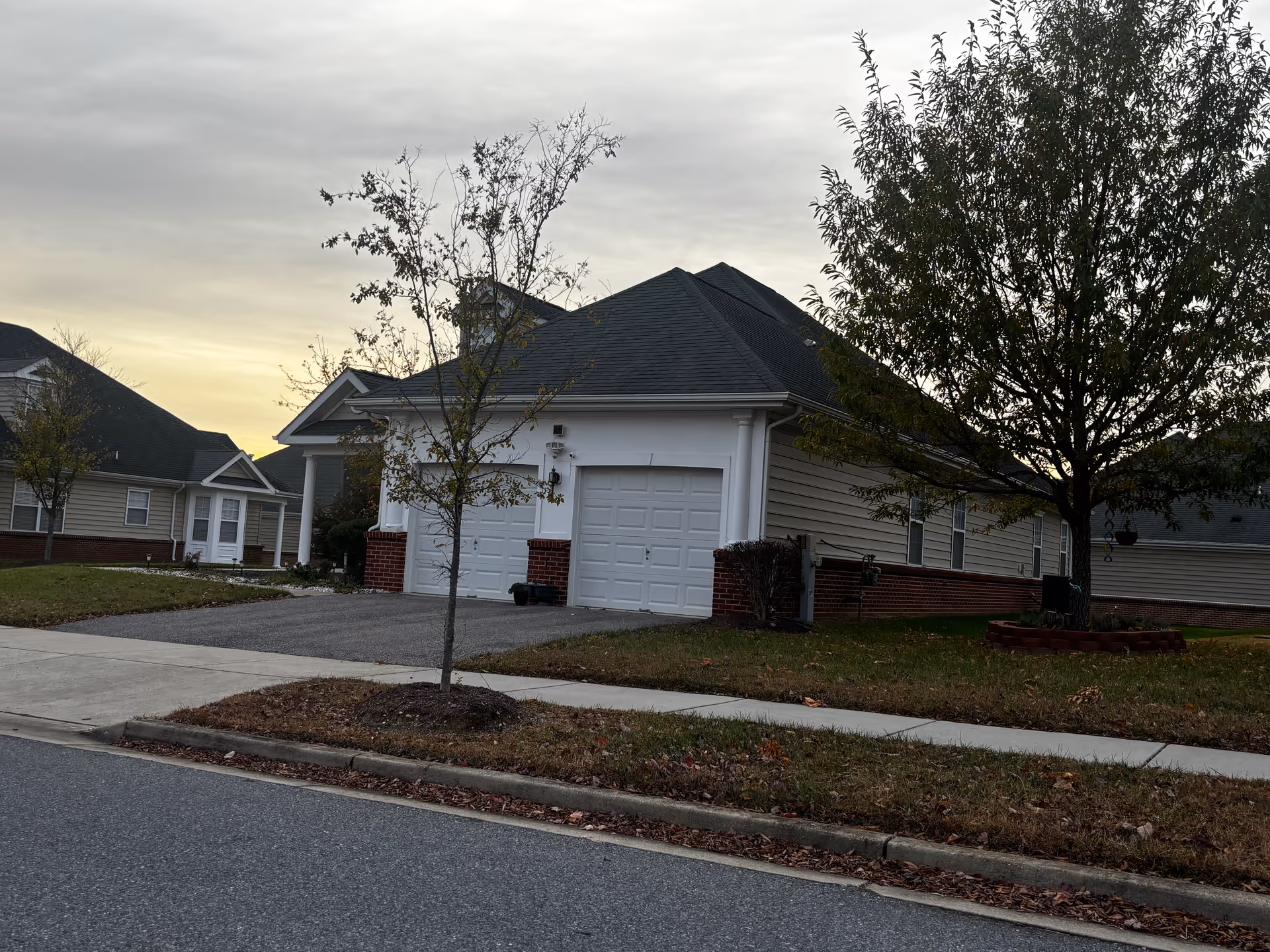 Exterior view of a residential building with a double garage door, a small tree in the front yard, and a sidewalk along the street. The building has beige siding with a brick base and a dark shingled roof. The sky is overcast with a hint of sunset light.