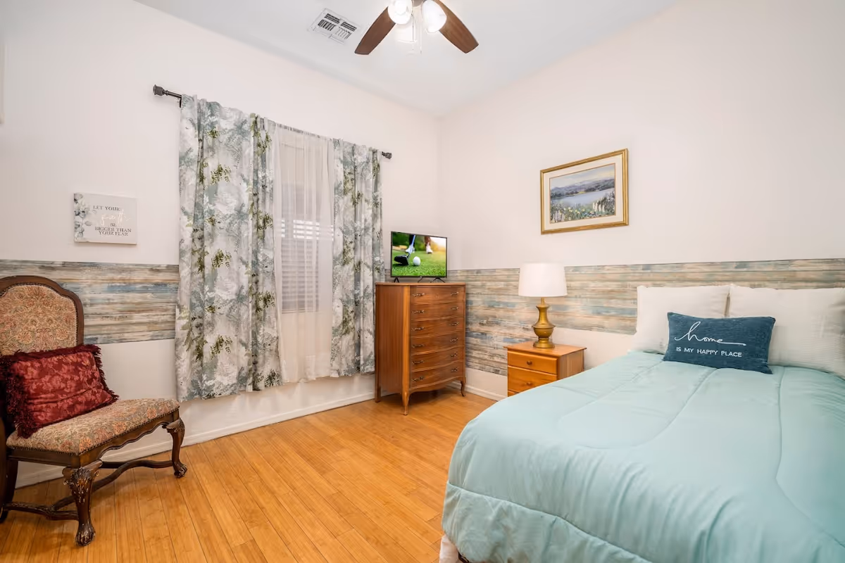 A cozy bedroom with a light blue bedspread and a decorative pillow that reads 'home is my happy place.' The room features a wooden chair with a red cushion, a wooden dresser with a small TV on top, a wooden nightstand with a lamp, floral curtains on the window, and a framed landscape painting on the wall.