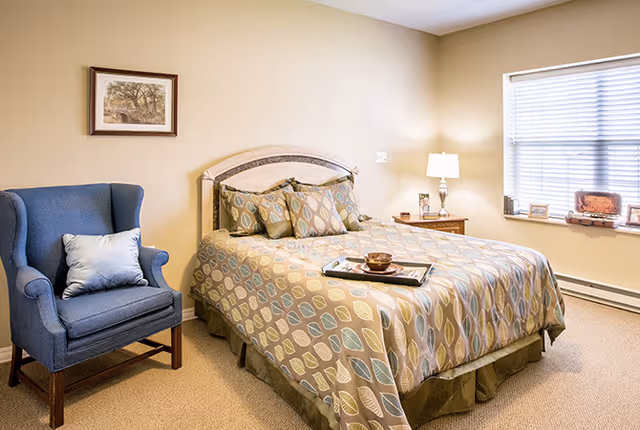 A cozy bedroom with a bed covered in a patterned bedspread featuring leaf designs in muted colors. There are several matching pillows on the bed and a tray with a cup and bowl placed on it. To the left of the bed is a blue upholstered armchair with a light-colored pillow. A framed picture hangs on the beige wall above the chair. On the right side of the bed is a wooden nightstand with a lamp, a small plant, and a few decorative items. A window with blinds lets in natural light on the right wall.
