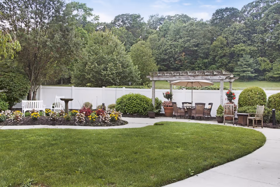 Outdoor landscaped courtyard with a pergola-covered dining area, seating, flowerbeds, and a grassy lawn.