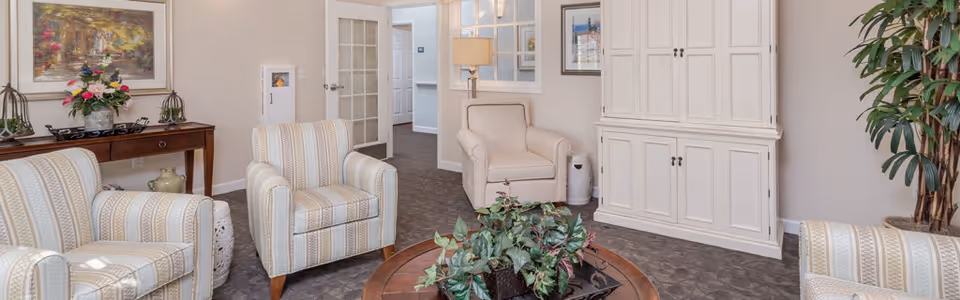 A cozy living room area with striped armchairs arranged around a round wooden coffee table with a plant centerpiece. There is a white cabinet against the wall, a side table with a lamp, a framed painting, and a potted plant in the corner. The room has neutral tones and a carpeted floor.