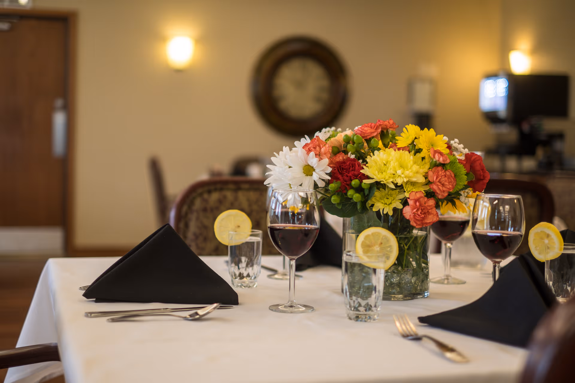 A dining table set with black folded napkins, glasses of water with lemon slices, and glasses of red wine. A colorful bouquet of flowers in a glass vase is placed in the center of the table. The background shows a wall clock and some furniture in a softly lit room.