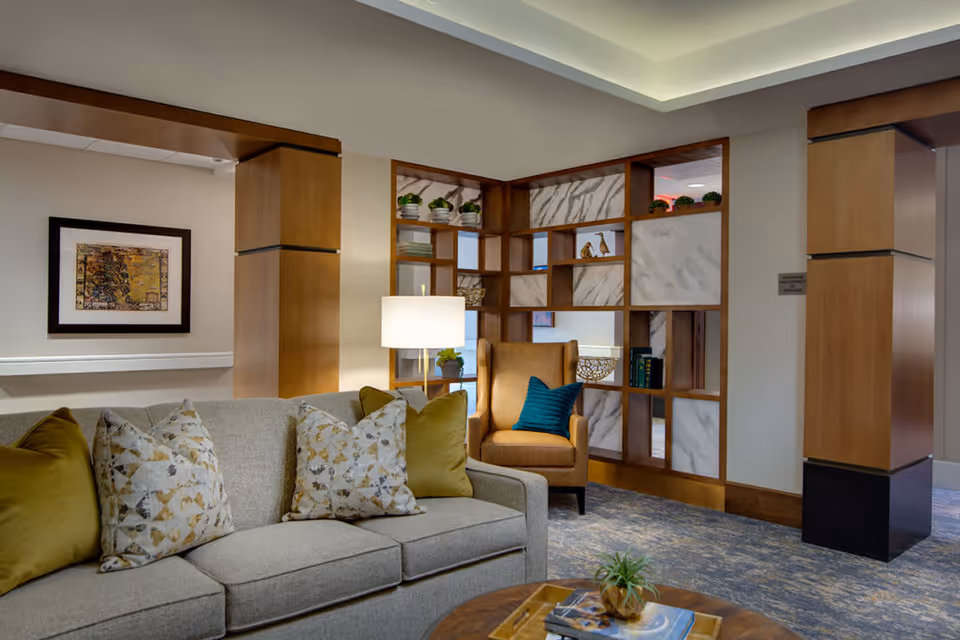 A cozy living room area featuring a light gray sofa with patterned and solid green pillows, a tan armchair with a blue pillow, a wooden coffee table with a small plant and books, a floor lamp, and a decorative wooden shelving unit with plants and ornaments. The room has warm lighting and a carpeted floor.