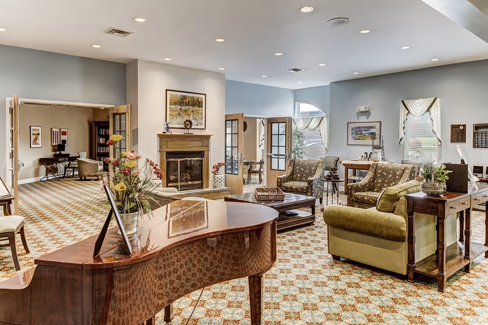 Bright, spacious senior living common room with a grand piano, seating area, fireplace, and patterned carpet.