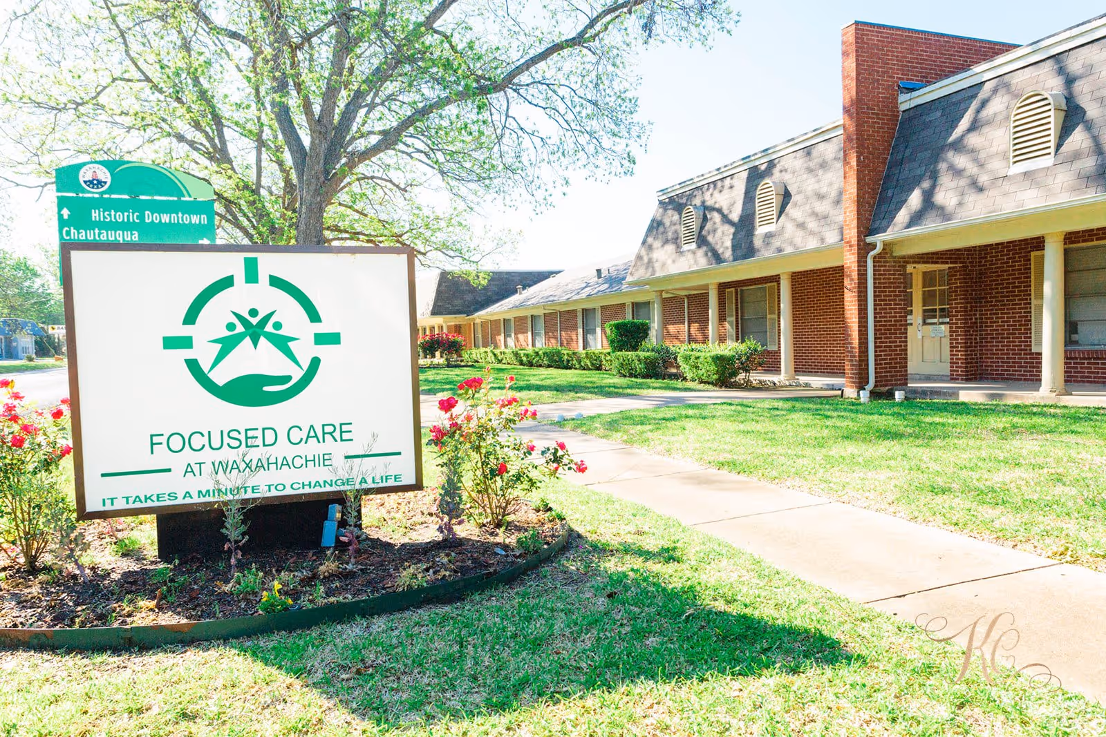 Exterior view of Focused Care at Waxahachie, showing a brick building with a sidewalk and green lawn. A large sign in the foreground displays the facility's name and logo, with flowers planted around the sign. A green street sign in the background points to Historic Downtown Chautauqua.