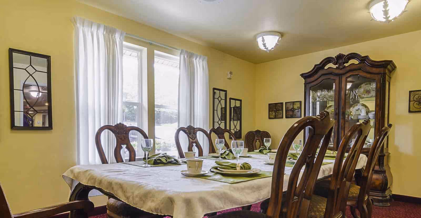 A formal dining room with a long table covered with a white tablecloth, set with green napkins, plates, cups, and glasses. The room has yellow walls, a large window with white curtains, wooden chairs with ornate backs, and a wooden china cabinet displaying dishes and glassware.