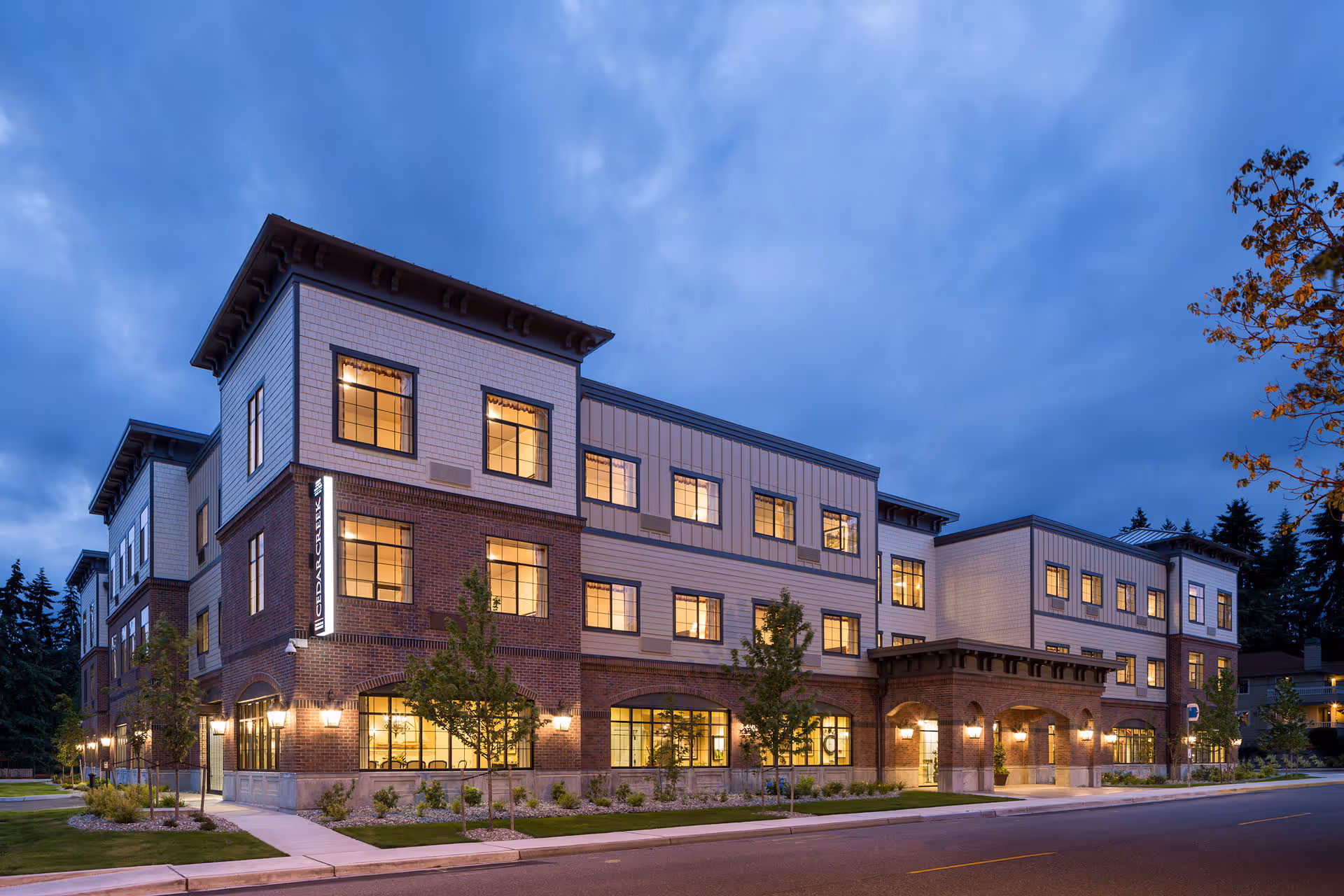 Exterior view of a three-story building at dusk with lights on inside. The building has a combination of brick and light-colored siding with multiple windows and a covered entrance. Trees and landscaping are visible along the sidewalk in front of the building.