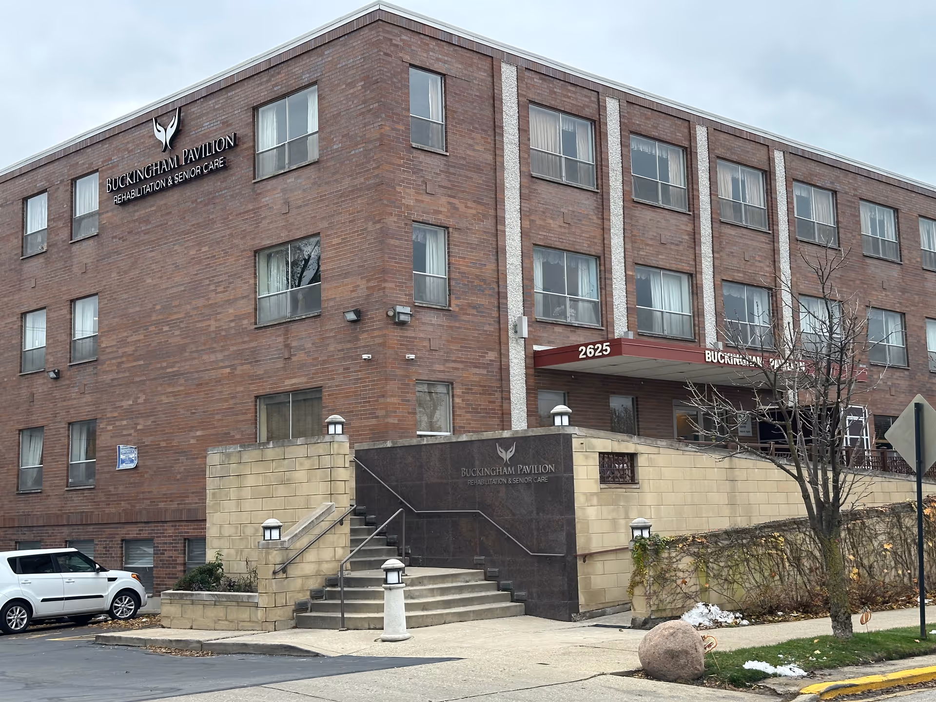Exterior view of Buckingham Pavilion Rehabilitation & Senior Care building, a three-story brick structure with multiple windows, a staircase leading to the entrance, and a sign displaying the facility's name and address 2625.