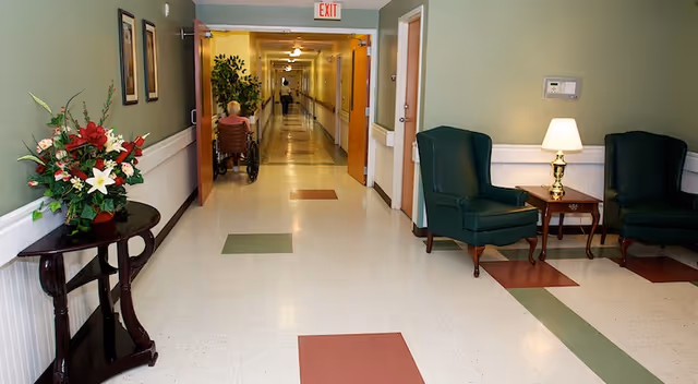 A hallway in a senior living facility with a person in a wheelchair moving down the corridor. The hallway has green and red square tiles on the floor, light green walls, and handrails on both sides. There are two green armchairs and a small table with a lamp on the right side, and a table with a floral arrangement on the left side near the entrance to the hallway.