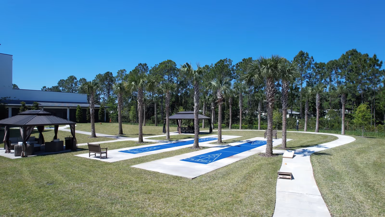 Outdoor recreational area at Paradise Pointe Assisted Living & Memory Care featuring two shuffleboard courts surrounded by palm trees, a curved concrete walkway, and two shaded seating gazebos on a sunny day with a clear blue sky.