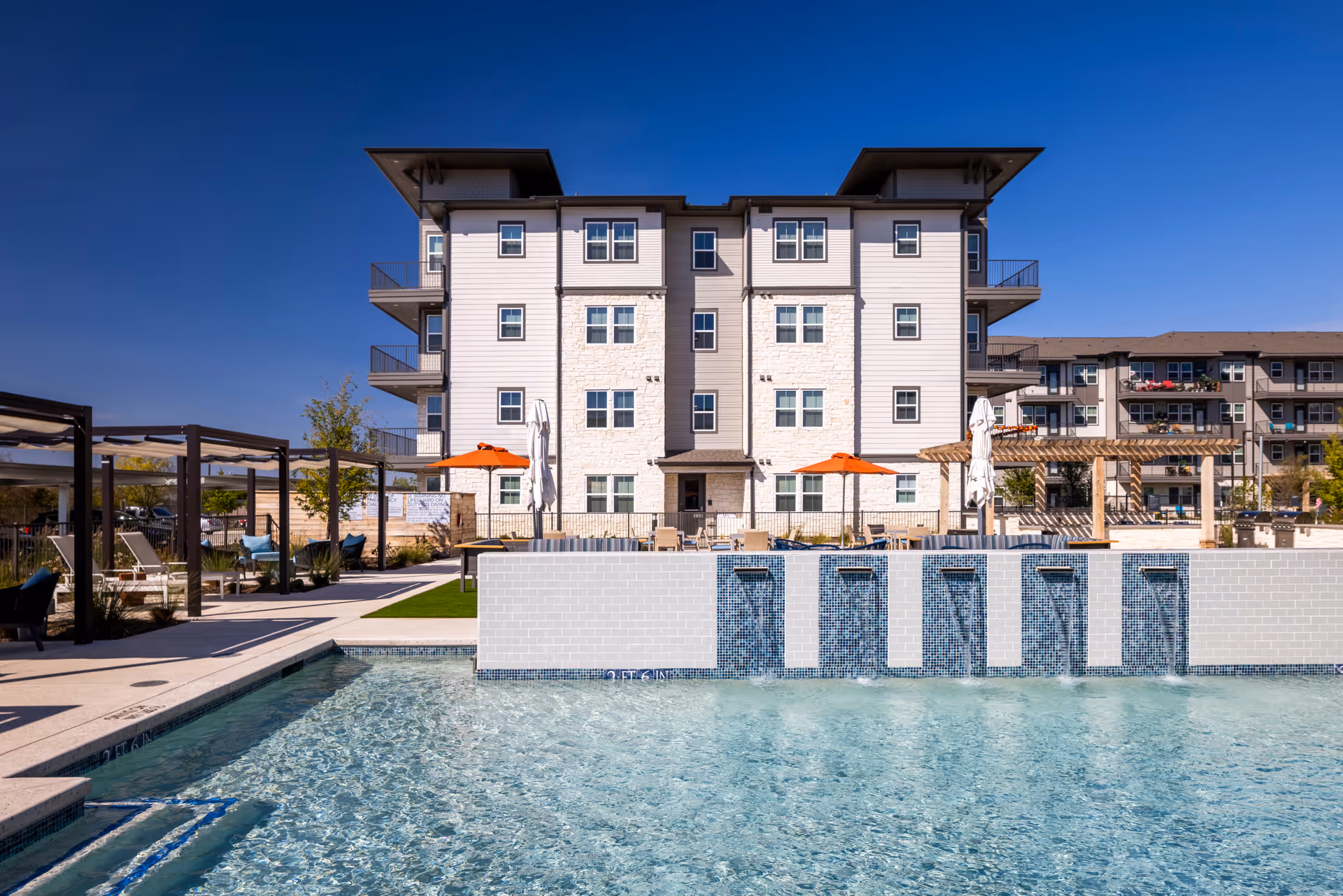Outdoor view of a senior living facility named Amberlin at The Station featuring a clear blue swimming pool with water fountains, lounge chairs under pergolas, orange umbrellas, and a multi-story residential building in the background under a clear blue sky.