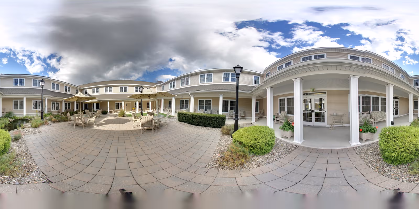 Outdoor courtyard area of Peregrine Senior Living at Delmar Place featuring paved walkways, patio tables with umbrellas, chairs, landscaped bushes, and a covered porch entrance to the building under a partly cloudy sky.