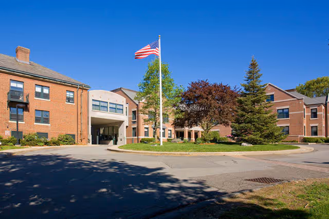 Exterior view of a brick senior living facility with an American flag on a flagpole in front, surrounded by trees and a circular driveway under a clear blue sky.