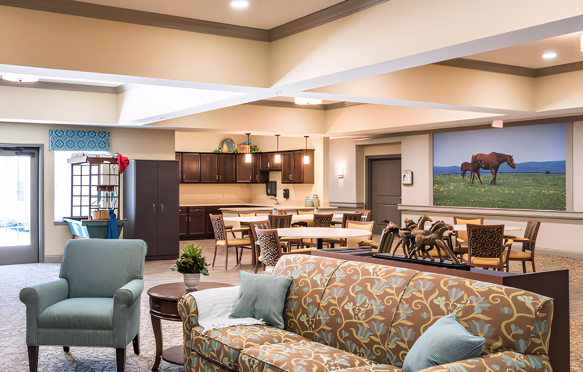 A spacious senior living common area with a patterned sofa and a light blue armchair in the foreground. Behind them are round tables with chairs, a kitchenette with dark wood cabinets, and a large wall art featuring horses in a green field. The room has beige walls and ceiling with recessed lighting.