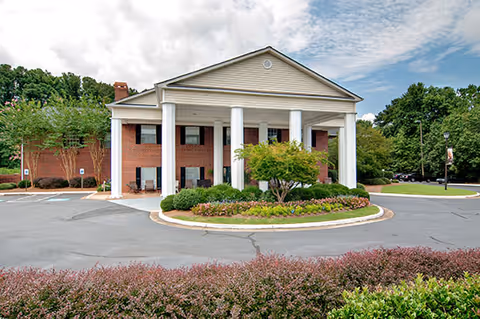 Front exterior view of a brick building with white columns and a triangular pediment, surrounded by landscaped greenery and a circular driveway under a partly cloudy sky.