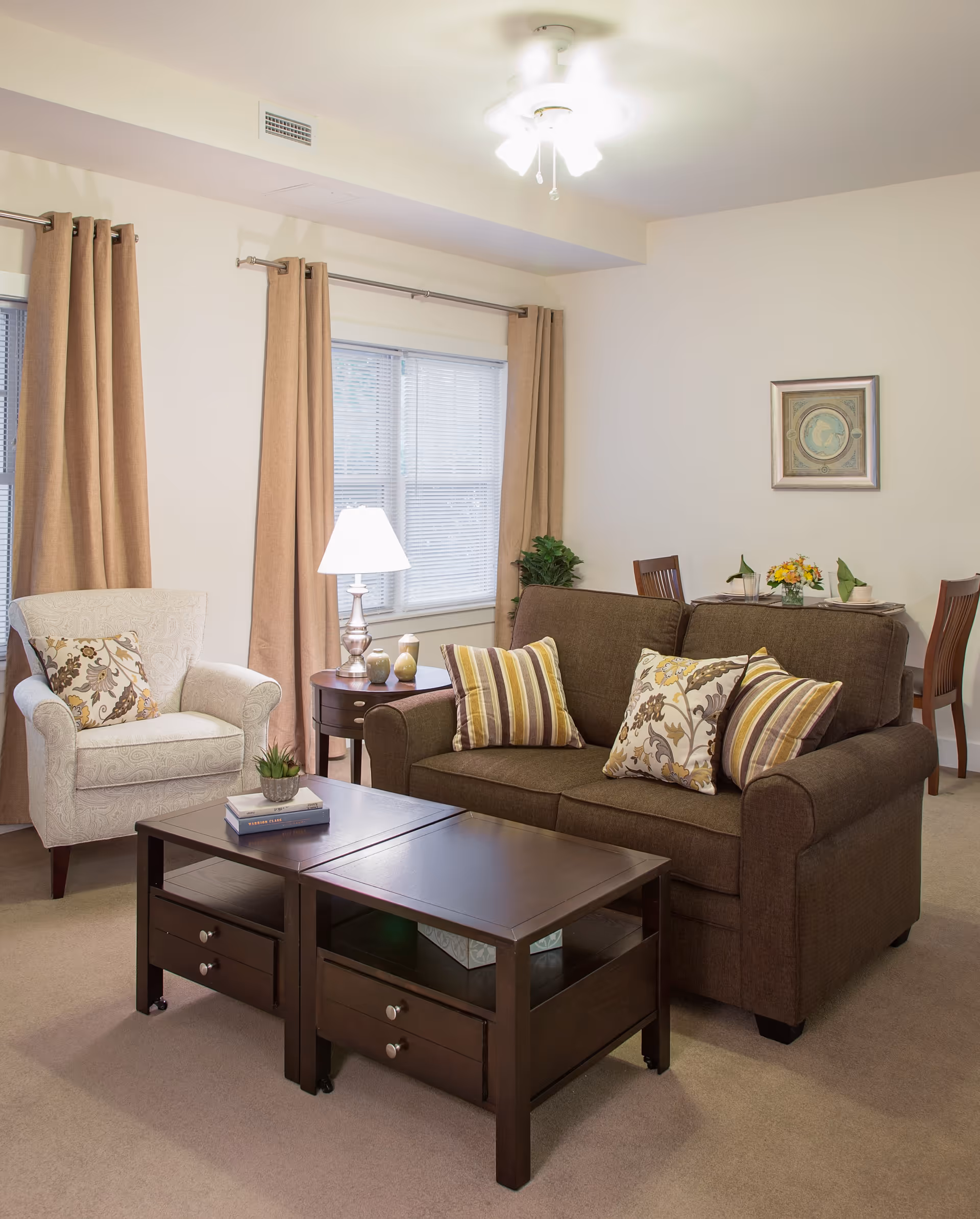 A cozy living room with a brown sofa adorned with three decorative pillows, a beige armchair with a patterned pillow, and a dark wooden coffee table with books and a small plant on it. Behind the sofa is a small dining table set with plates, glasses, and a flower arrangement. The room has beige walls, beige curtains on the windows, a ceiling fan with lights, and a framed artwork on the wall.