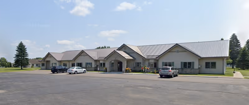Single-story assisted living building with a central entrance, parking lot and several cars under a clear sky.