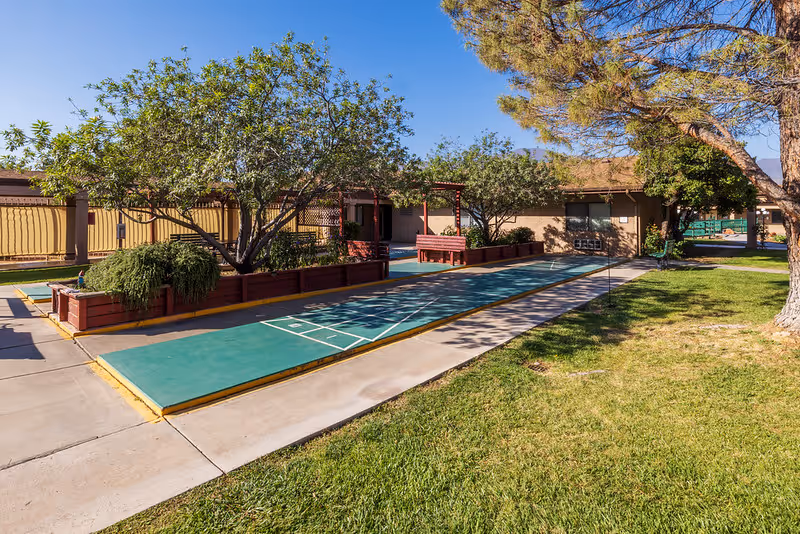 Outdoor courtyard featuring shuffleboard courts, raised planter boxes, trees, benches and nearby single-story buildings under a blue sky.
