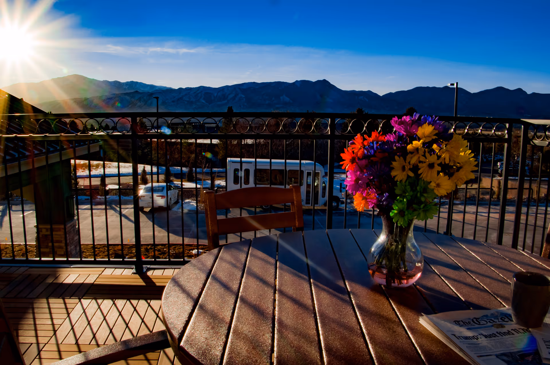 Sunlit balcony patio with a table and vase of colorful flowers overlooking a parking lot and distant mountains.