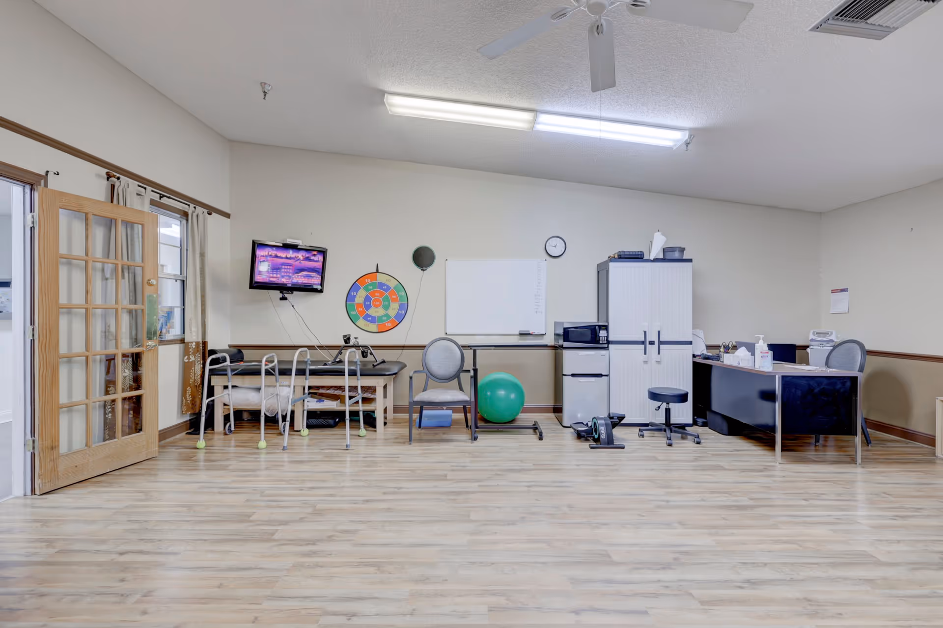 A spacious room with light wood flooring and beige walls, featuring a therapy or exercise area with a walker, exercise ball, and a therapy table. There is a wall-mounted TV, a colorful target board, a whiteboard, a clock, and a desk with office supplies. The room has a ceiling fan and fluorescent lighting, with a wooden door with glass panels on the left side.