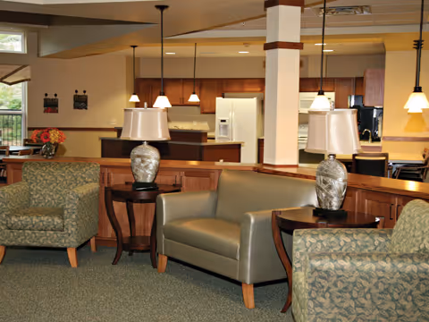 Interior view of a senior living facility common area with a seating arrangement including two patterned armchairs and a gray leather loveseat. There are two side tables with decorative lamps on either side of the loveseat. In the background, a kitchen area with wooden cabinets, a refrigerator, and pendant lights is visible.