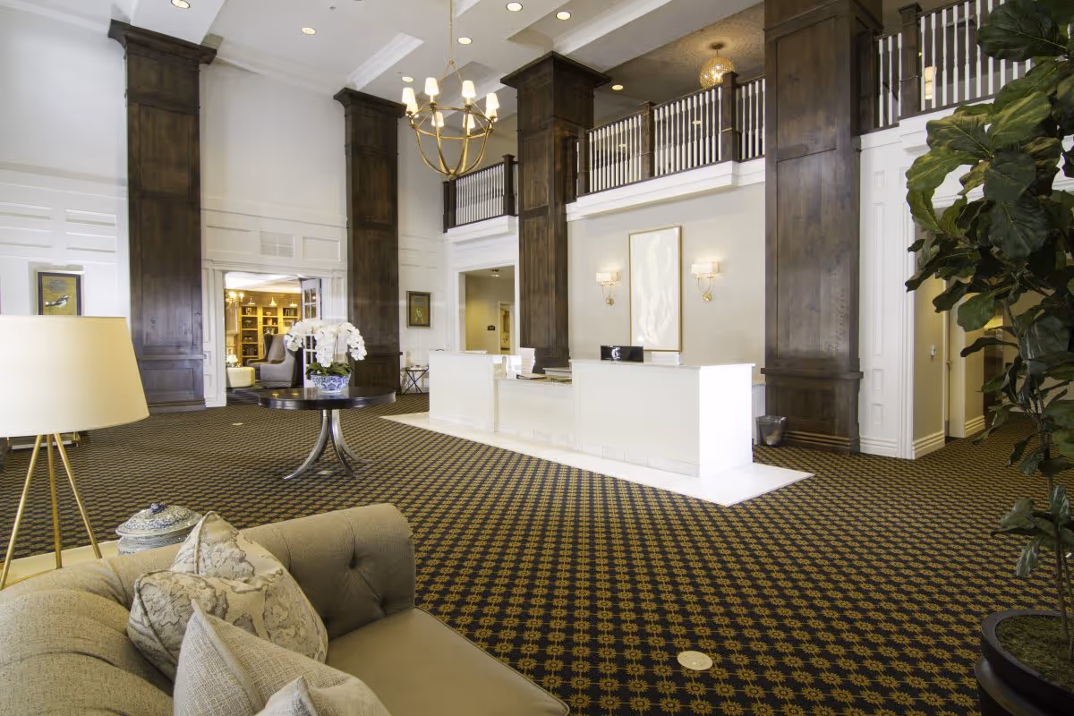 Spacious and elegant lobby area with a patterned carpet, large dark wooden columns, a white reception desk, a round table with a flower arrangement, a beige sofa with decorative pillows, a floor lamp, and a large plant. The ceiling is high with a chandelier and recessed lighting.