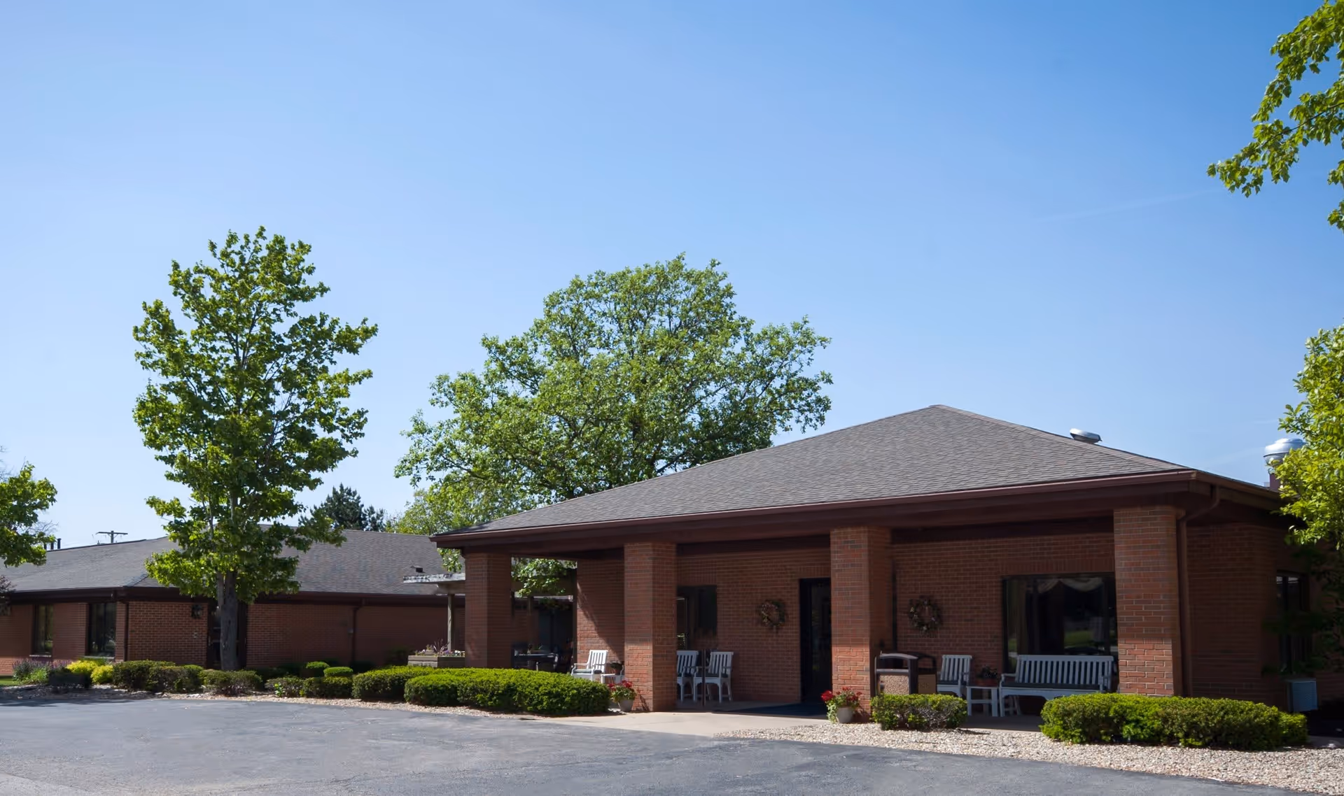 Exterior view of a single-story brick building with a covered entrance supported by brick columns. There are white benches and chairs on the porch, surrounded by neatly trimmed bushes and trees under a clear blue sky.