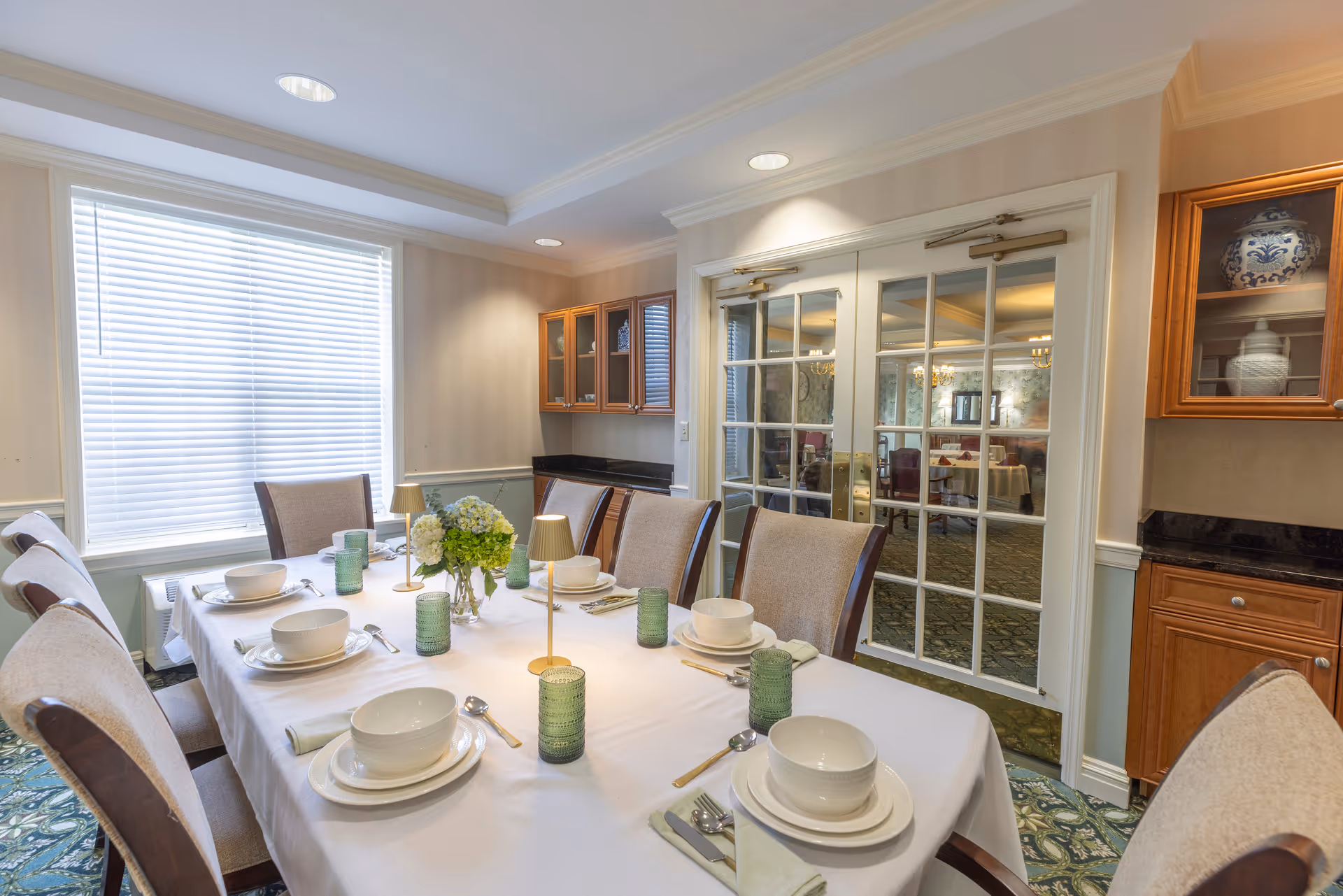A well-lit dining room with a rectangular table set for six people. The table is covered with a white tablecloth and set with white bowls, plates, silverware, green textured drinking glasses, and small gold lamps. A vase with green and white flowers is in the center of the table. The room has beige walls, a large window with white blinds, wooden cabinets with glass doors, and double glass doors leading to another dining area.