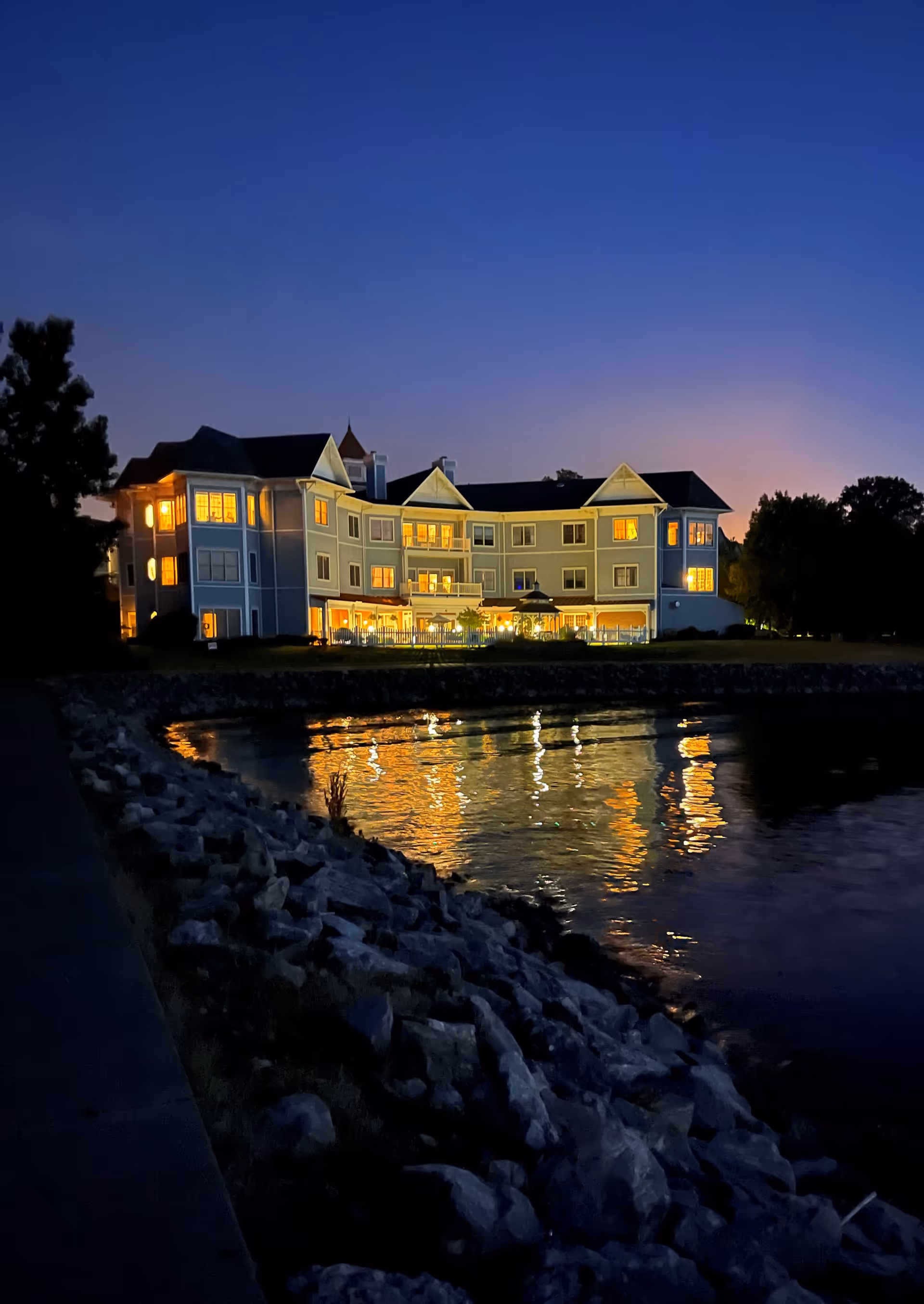A lit senior living building on the water at dusk with its lights reflecting on the surface and a rocky shoreline in the foreground.