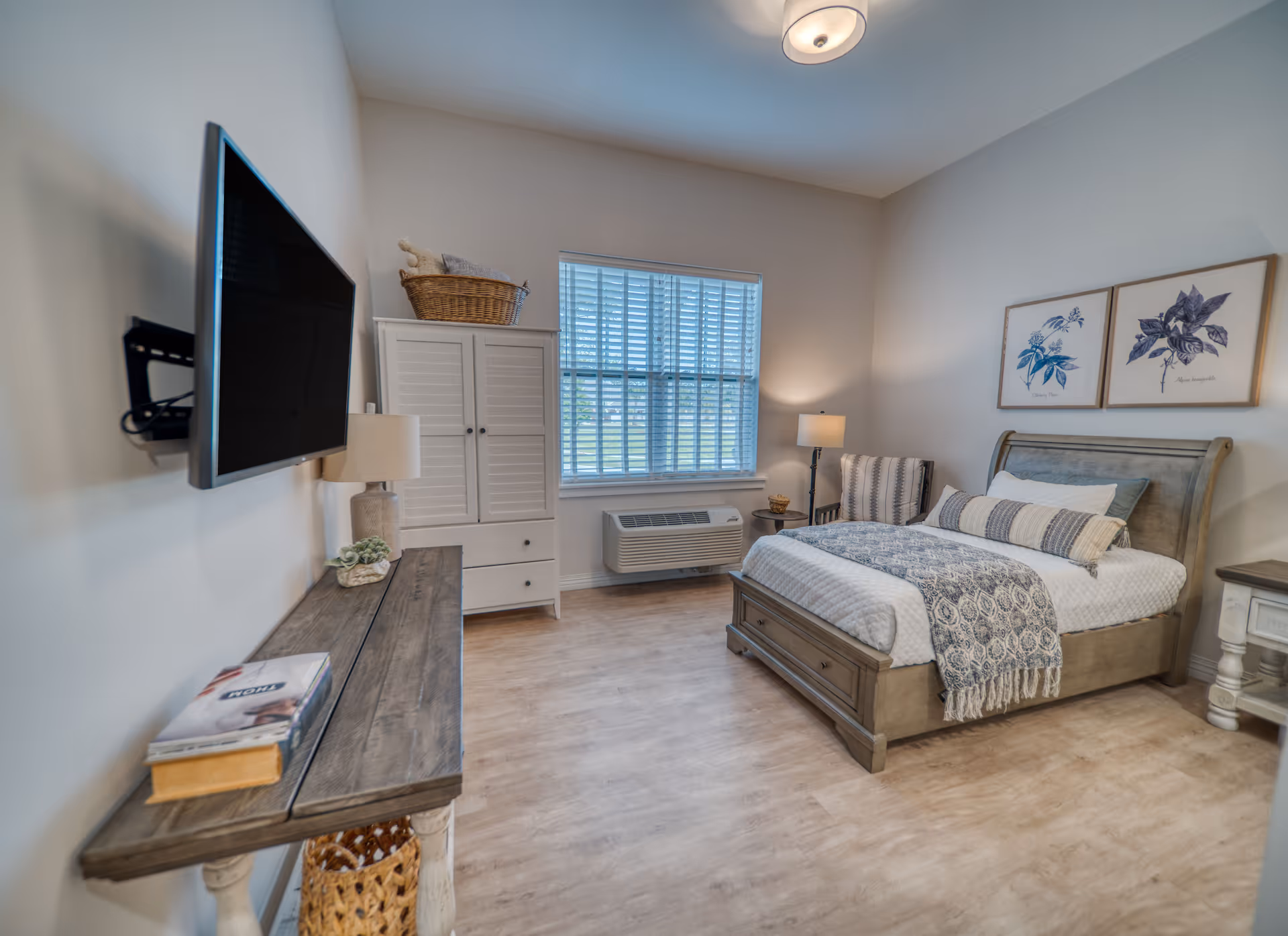 A cozy bedroom in an assisted living facility featuring a wooden bed with white and gray bedding, two framed botanical prints on the wall above the bed, a wooden nightstand with a lamp, a cushioned chair with a striped pillow, a window with blinds, a white cabinet with a basket on top, a wooden console table with books and a lamp, and a wall-mounted flat-screen TV.