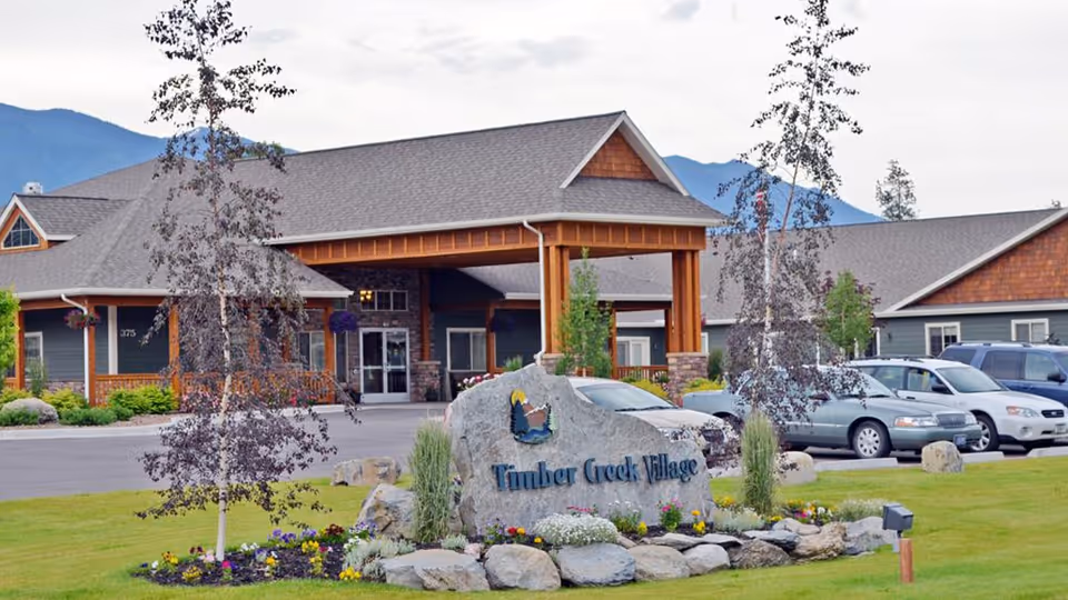 Exterior view of Timber Creek Village Community building with a large stone sign in front surrounded by landscaping and parked cars. The building has a covered entrance with wooden beams and a mountain range visible in the background.